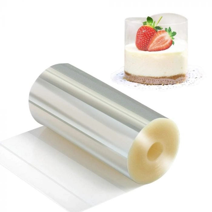 A cylindrical roll of clear plastic material, known as a cake collar, is positioned on a white surface, with one end unrolled. Next to it, there is a small, heart-shaped cheesecake topped with a sliced strawberry and a green leaf, displayed in a transparent container. The dessert sits on a decorative white doily.