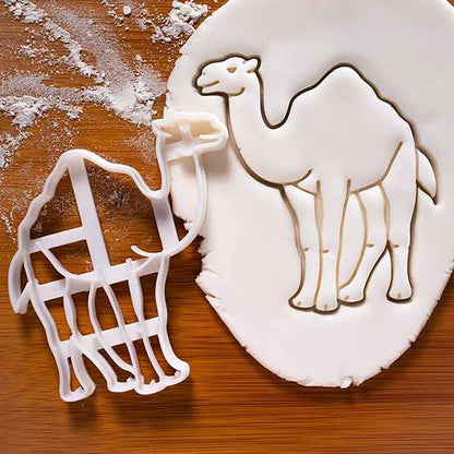 A camel-shaped cookie cutter is positioned next to an impression of a camel cut out of white fondant. The background shows a wooden surface sprinkled with flour. The cutter is white plastic, and the fondant has a detailed outline of a camel.