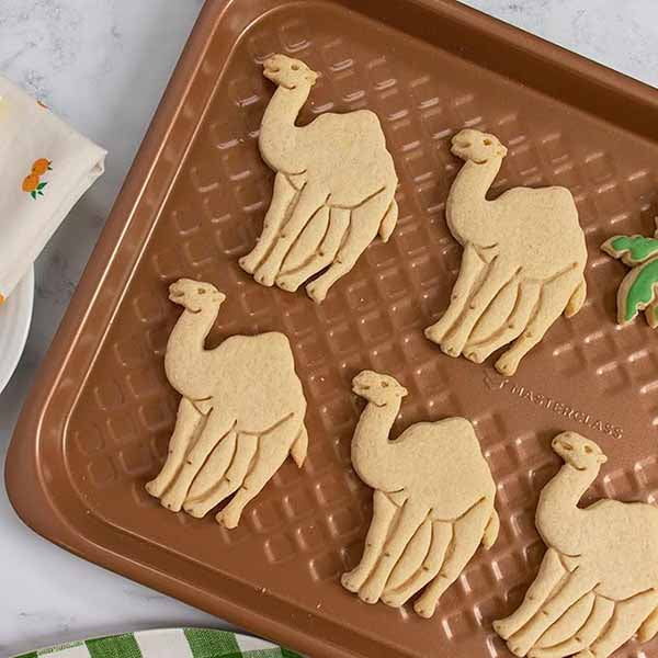 A baking tray with five camel-shaped cookies arranged on it. The cookies are light beige and appear freshly cut, with a simple design representing camels. A green leaf-shaped cookie is also visible on the tray. In the background, a patterned napkin can be seen beside the tray.