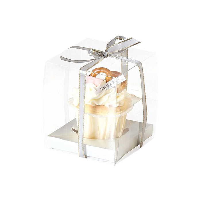 A clear PVC box containing a single cupcake is tied with a silver ribbon. The cupcake features a light-colored base with decorative frosting in shades of pink and white, and is topped with small colorful items resembling candy. The box has a square shape and rests on a flat, white base. A small label on the box displays the word "SUGAR."