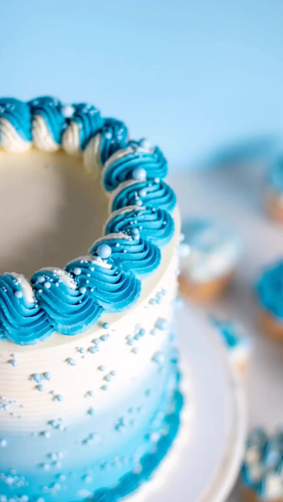 A beautifully decorated cake featuring a gradient of blue and white frosting. The top edge is adorned with a wave pattern of bright blue icing, with small white and light blue decorative pearls scattered across the cake&