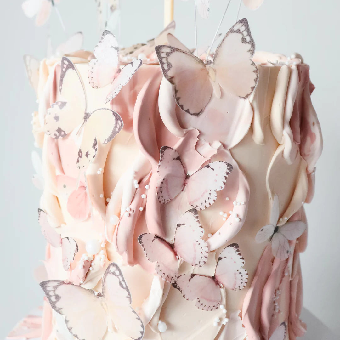 An alt text description for the image could be:
"A beautifully decorated cake covered with layers of creamy frosting in shades of pink and white. The cake is adorned with numerous delicate butterfly decorations, featuring soft pink hues and intricate details on their wings. Small white beads complement the design, enhancing the overall elegance of the cake."