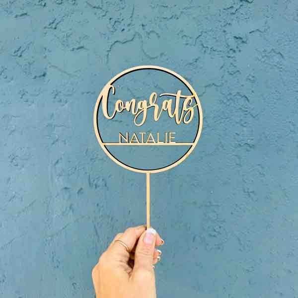 A gold acrylic cake topper being held by a hand against a textured blue wall. The topper features the words &quot;Congrats&quot; in a cursive font and &quot;NATALIE&quot; in a bold, uppercase font, enclosed in a circular design.