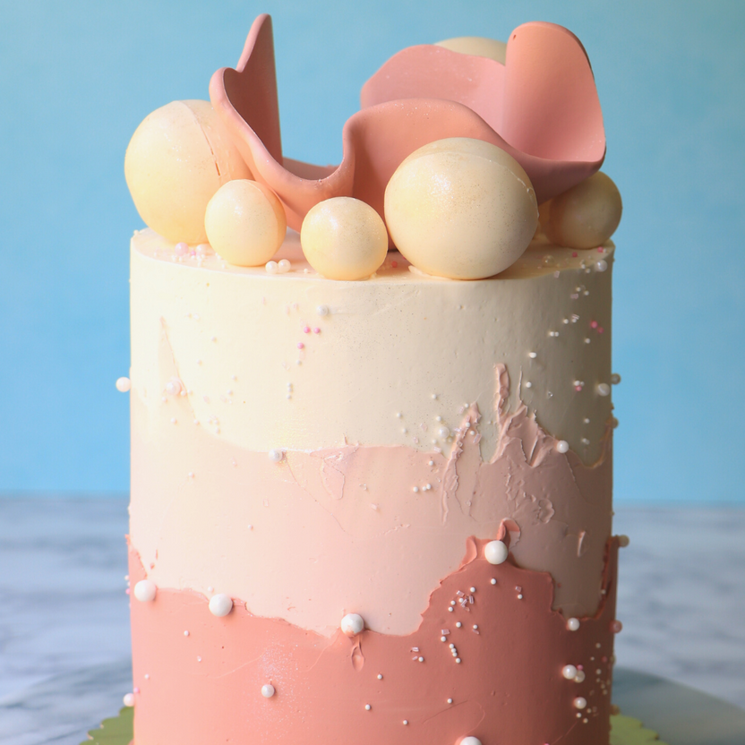 A visually appealing cake with a smooth, layered frosting in shades of pale yellow, soft pink, and deeper pink. It is topped with decorative elements, including round, glossy balls in various sizes, and a pink, wavy piece of fondant. Small, white sugar pearls are scattered across the surface, enhancing the cake&