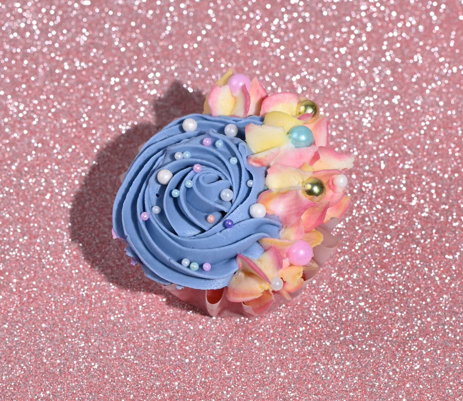 A cupcake is decorated with swirls of blue frosting and adorned with colorful edible pearls and pastel flower decorations. The cupcake is placed on a sparkly pink background, enhancing its vibrant colors.