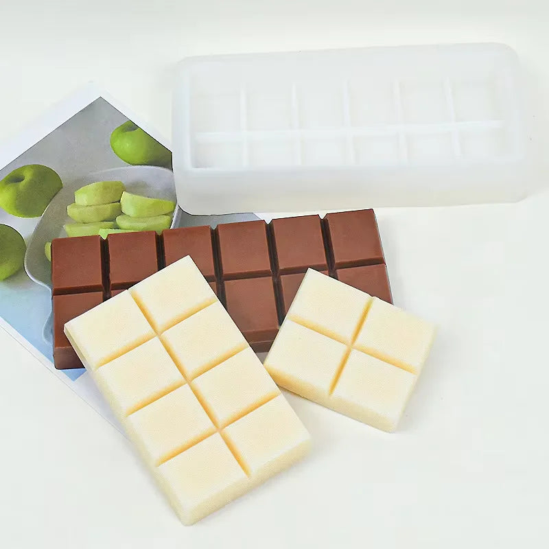 An image featuring a silicone chocolate bar mold placed above two sets of chocolate pieces. The mold is white and rectangular, with eight square cavities. Surrounding the mold are several chocolate pieces: two are light in color, resembling white chocolate, and six are darker, resembling milk chocolate. In the background, there is a photo of green apples and a plate. The overall composition is set against a light background, focusing on the chocolate and mold.