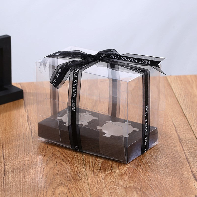 A clear plastic cupcake box designed to hold two portions. The box is decorated with a black ribbon that reads &quot;BEST WISHES FOR YOU.&quot; It sits on a wooden surface, with a partial black stand visible in the background.