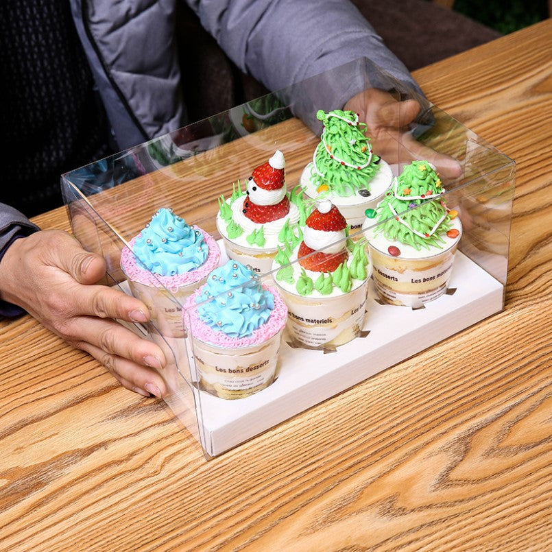 A clear cupcake box containing six decorated cupcakes is held by a person. The cupcakes feature various colorful frosting designs, including blue, pink, and green, with decorations like strawberries and festive sprinkles. The box is placed on a wooden table, showcasing its transparent sides.