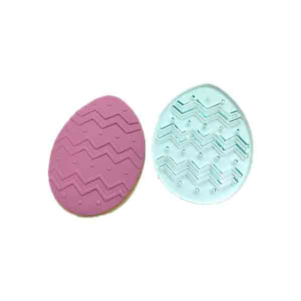 Two egg-shaped cookie stamps are displayed side by side. The left stamp is pink with a zigzag pattern and small raised dots. The right stamp is translucent blue with a similar zigzag design and raised lines. Both stamps are designed for embossing fondant, particularly for Easter-themed baking.