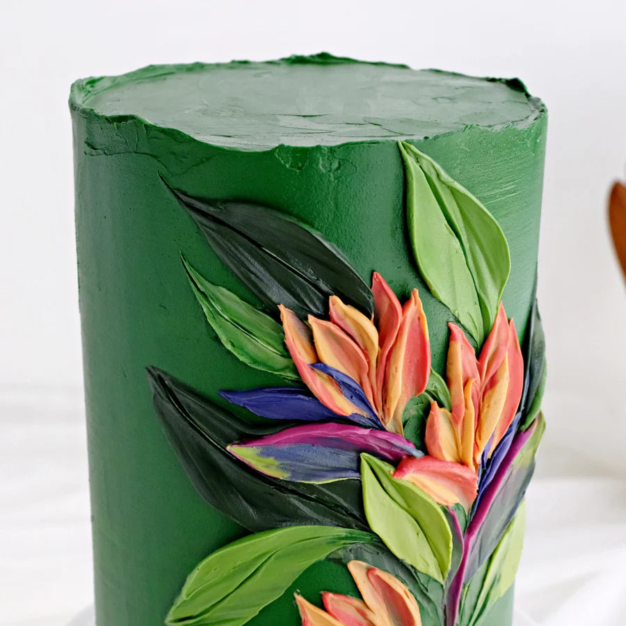 The image features a tall, round cake covered in a smooth green frosting. The cake is decorated with vibrant, three-dimensional floral designs featuring shades of pink, orange, blue, and yellow, alongside green leaves. The design creates a lively, tropical appearance against the green backdrop of the cake. The background is light and slightly blurred, emphasizing the cake&