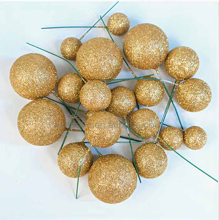 A collection of gold glitter ball cake toppers arranged on a white surface. The set includes various sizes of spherical balls, all coated in gold glitter, with some attached to thin green stems.