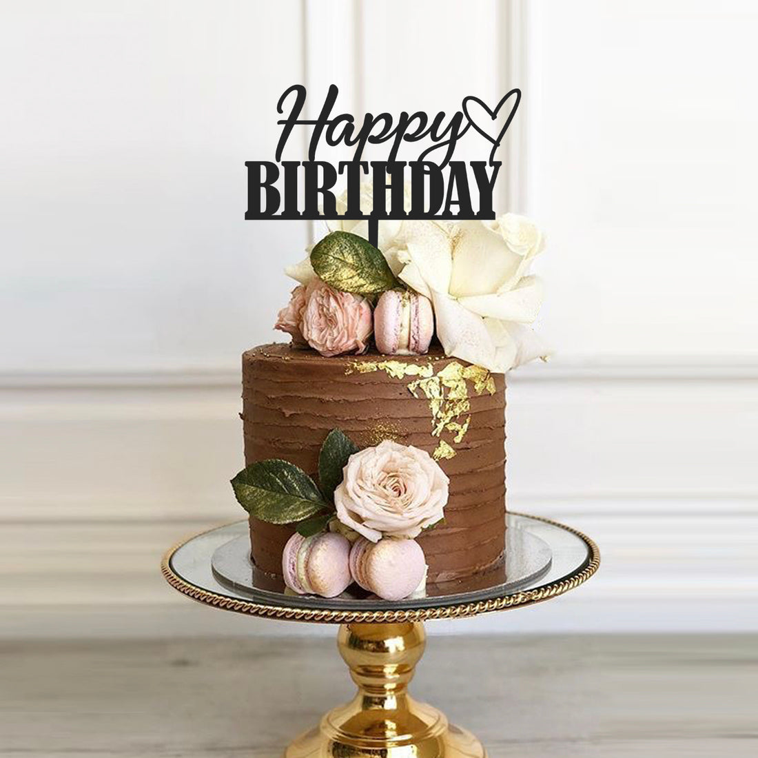 A beautifully decorated chocolate birthday cake topped with a black "Happy Birthday" cake topper featuring a heart design. The cake is adorned with fresh flowers, including white and pink roses, and includes pale pink macarons. It is displayed on a round, mirrored cake stand with a gold base, set against a light background.