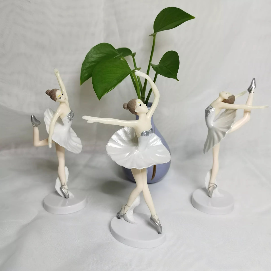 Three figurines of ballerina dancers in white dresses and silver accents are arranged on a light background. The figures are posed in different dance positions: one with a raised arm and a pointed foot, another with an outstretched arm and a leg raised high, and the third is poised on one leg with arms extended. A small vase with green leaves is visible in the background, adding a touch of greenery to the scene.