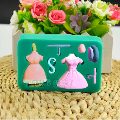 A green silicone mould featuring various shapes for crafting, including two dress silhouettes in pink shades, a hanger shape, and additional decorative elements like a bow and accessories. The mould is placed on a woven surface with a background of colorful faux flowers.