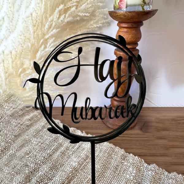 The image features a black acrylic cake topper that reads &quot;Hajj Mubarak&quot; in elegant, cursive script. The text is enclosed within a circular design adorned with small leaves, giving it a decorative look. The cake topper is situated in a softly lit environment, highlighting its design against a neutral background. A textured cloth and wooden stand are visible in the setting.