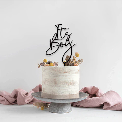 A round, two-tiered cake with a textured cream-colored frosting is decorated with dried flowers on top. A black acrylic cake topper that says &quot;It&