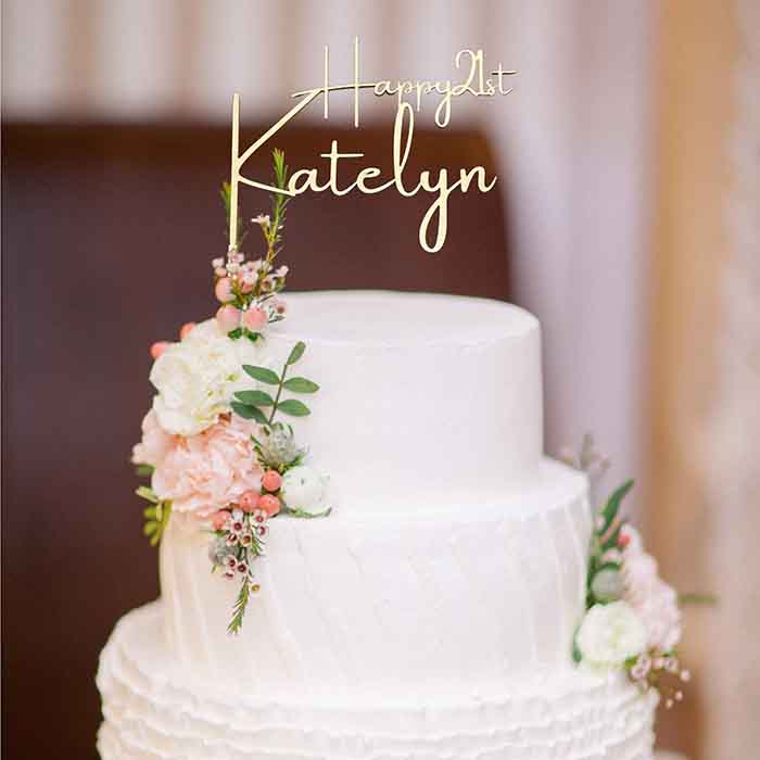 A two-tier white cake topped with a personalized acrylic cake topper that reads &quot;Happy 21st Katelyn.&quot; The cake is decorated with delicate pink and white flowers, along with green leaves, adding a touch of elegance to the design. The background is softly blurred, highlighting the cake.