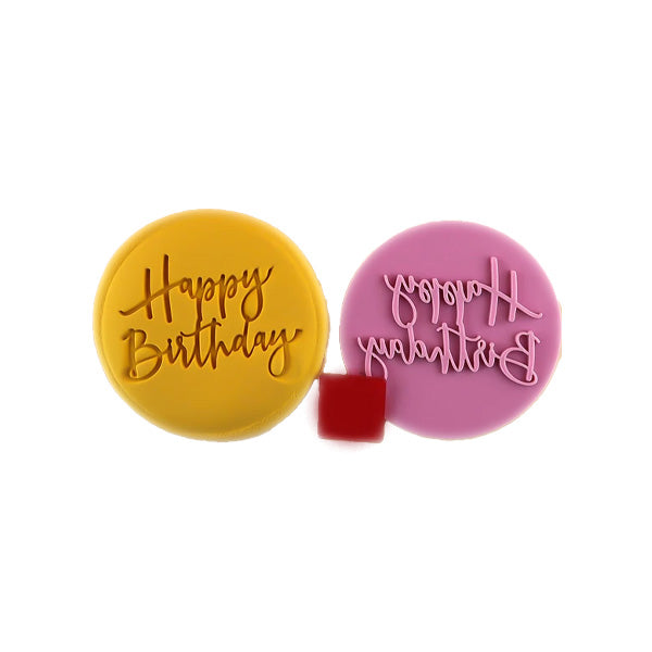 Two round cookie stamps for embossing fondant. One stamp is yellow and features the text &quot;Happy Birthday&quot; in a cursive font. The other stamp is pink and has the word &quot;Birthday&quot; in a mirrored cursive script. A small red square block is positioned between the two stamps.