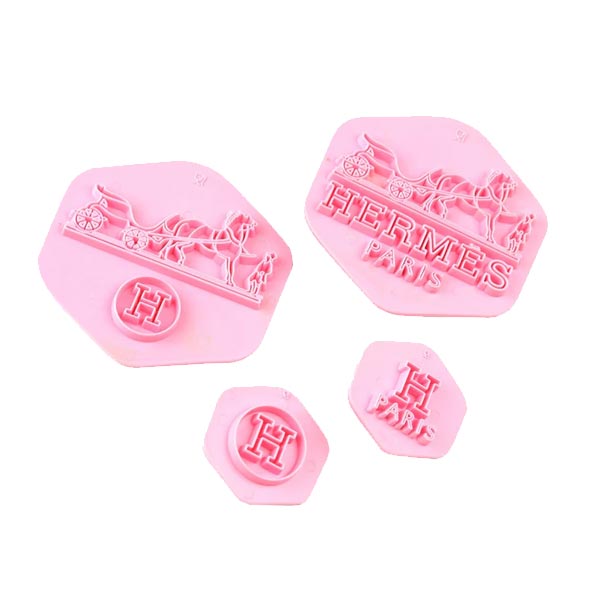 A set of four fondant embosser cutters in pink silicone, shaped in hexagons. The larger cutouts feature the word &quot;HERMES&quot; with &quot;PARIS&quot; underneath, along with a design of a horse and carriage. Two smaller cutouts display the letter &quot;H&quot; and the word &quot;PARIS&quot;. The items are designed for cake decorating.