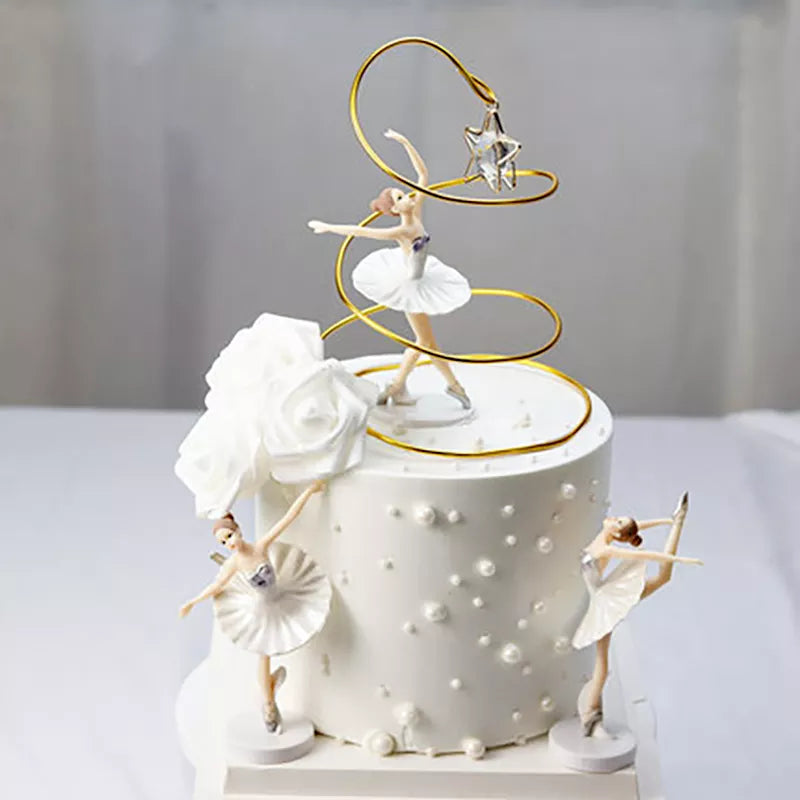 A decorative cake topper featuring three ballerina figurines on a white, round cake. The central ballerina is posed mid-dance with arms raised and is surrounded by a gold swirling decoration topped with a star. The cake is adorned with white roses and small white pearls, creating an elegant appearance suitable for a celebration.