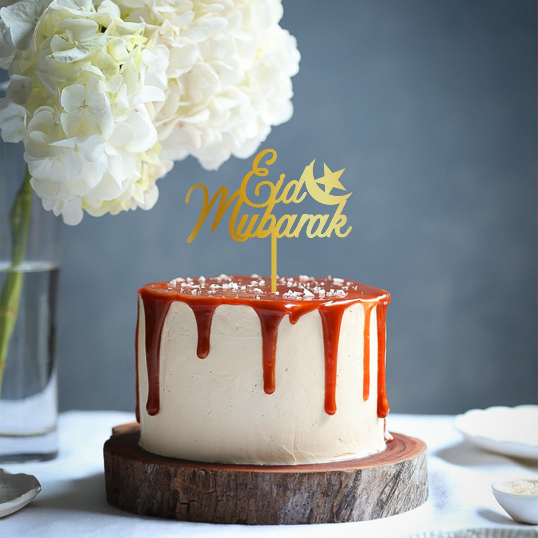 A decorated cake topped with a gold acrylic topper that reads &quot;Eid Mubarak&quot; accompanied by a crescent moon design. The cake has a smooth, light-colored frosting with a rich caramel glaze dripping down the sides, and is placed on a wooden base. In the background, there is a clear glass vase filled with white flowers.