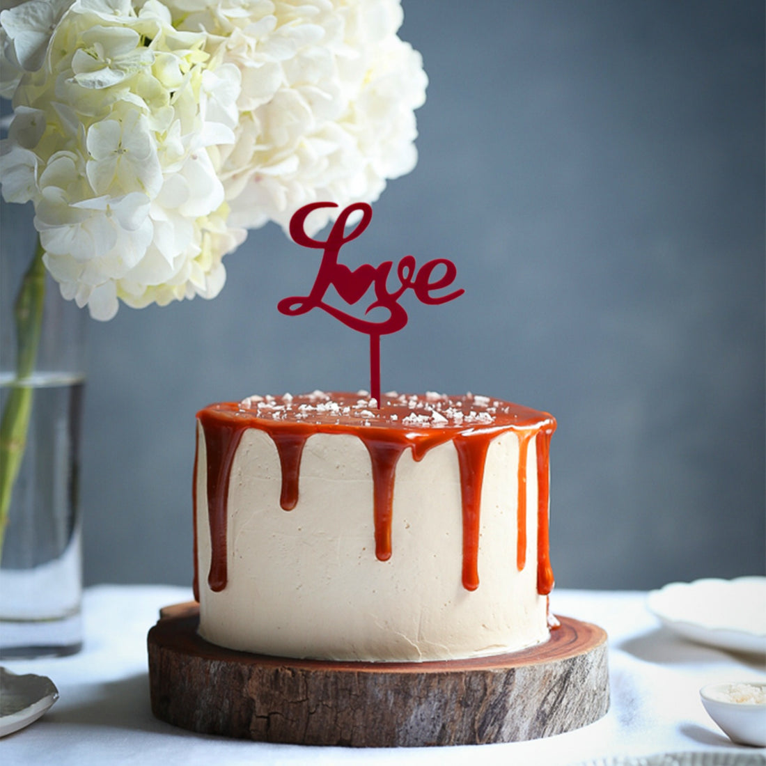 A round cake with creamy frosting is topped with a shiny caramel drizzle, which flows down the sides. Atop the cake is a red acrylic topper that reads &quot;Love&quot; in a decorative script. The cake is placed on a wooden serving board. Next to it, there is a glass vase containing white flowers, and a small plate with what appears to be dessert items. The background is softly blurred, highlighting the cake and its decorations.