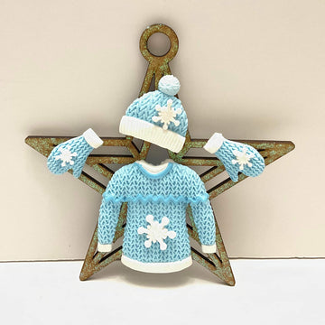 The image features a decorative star with a ring at the top. Positioned in front of the star is a blue knitted sweater adorned with a white snowflake design in the center. Above the sweater is a matching blue hat with a white pom-pom, and to the sides are two white and blue mittens. The overall theme emphasizes winter and holiday aesthetics.