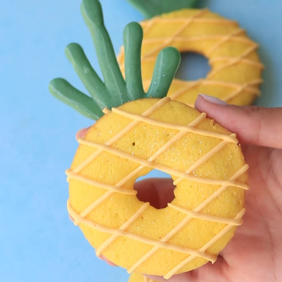 A person is holding a bright yellow donut shaped like a pineapple. The donut features a textured surface with a diamond pattern made of light brown icing and green icing for the pineapple leaves on top. In the background, there are additional similar pineapple-shaped donuts. The setting has a light blue surface.