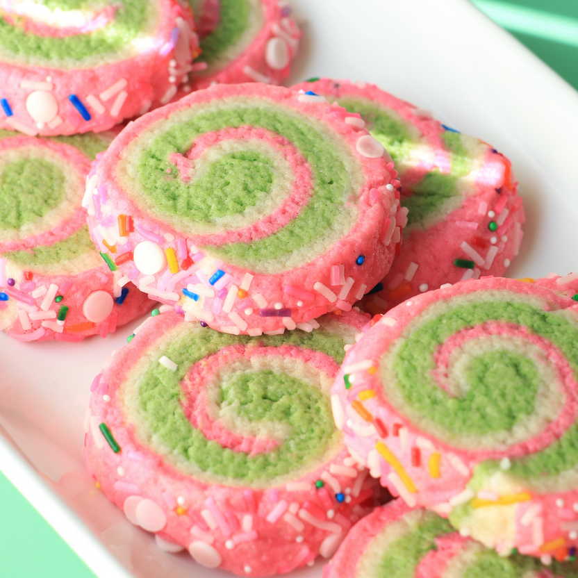 A close-up image of colorful, spiral-shaped cookies arranged on a white plate. The cookies feature a bright pink outer layer with a green swirl in the center, and are decorated with colorful sprinkles and small round candies. The background is a soft green, providing a vibrant contrast to the cookies.