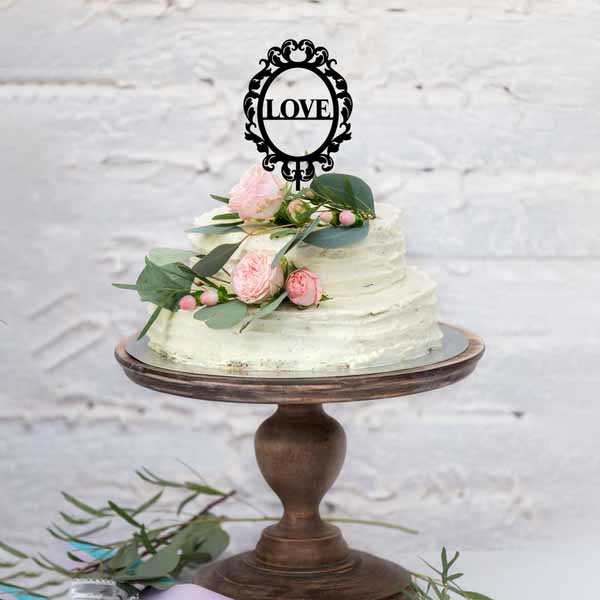 A three-tiered cake with a light green frosting is displayed on a wooden cake stand. The cake is adorned with pink roses, green leaves, and small berries. At the top of the cake, there is a black acrylic cake topper that features the word &quot;LOVE&quot; within an ornate frame. The background consists of a textured white wall.