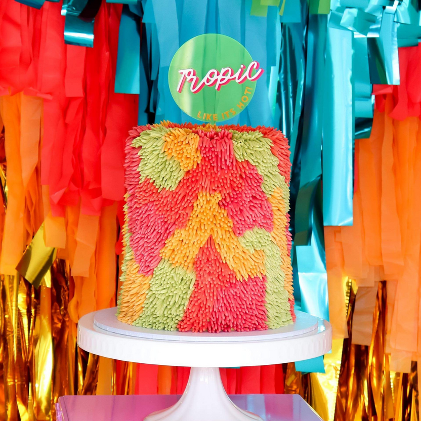 A colorful cake stands on a white pedestal, adorned with vibrant frosting in shades of pink, orange, and green, creating a textured floral appearance. Behind the cake, there is a festive backdrop of streamers in red, orange, blue, and gold. Prominently displayed on top of the cake is a circular sign that reads &quot;Tropic&quot; in pink lettering, accompanied by the phrase &quot;LIKE IT&