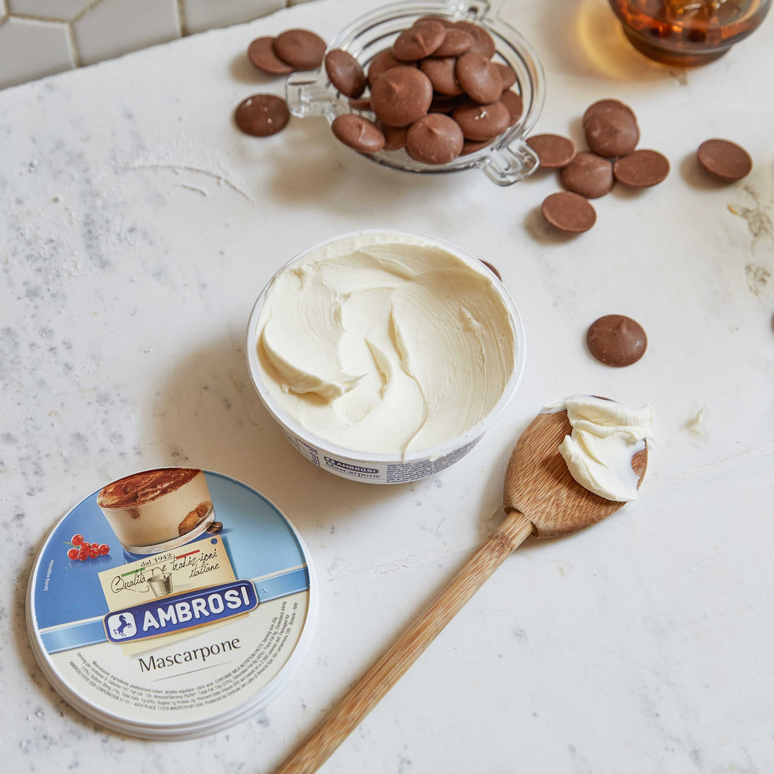 Flat lay of an open Ambrosi Mascarpone container on a marble surface, with its lid beside it. A wooden spoon with mascarpone rests nearby, surrounded by scattered chocolate pieces and a glass container filled with chocolates in the background.