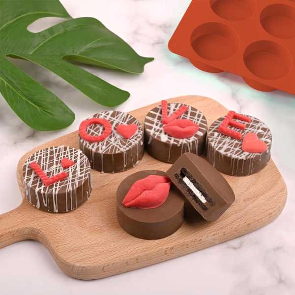 A wooden serving board displays a variety of chocolate mini cheesecakes. Some are topped with red fondant decorations shaped like lips and hearts, while others have the word "LOVE" made of red letters. A partially cut cheesecake reveals a filling inside. In the background, there is a green leaf and an orange silicone mold with 12 cavities. The scene has a light, festive feel.