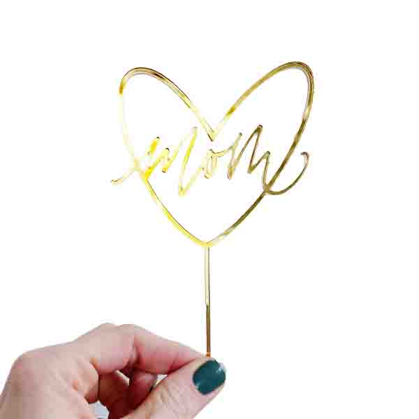 An image of a gold cake topper shaped like a heart with the word &quot;Mom&quot; elegantly scripted inside. A hand is holding the topper, which has a thin stem for inserting into a cake. The background is plain white, emphasizing the shiny gold color of the topper.