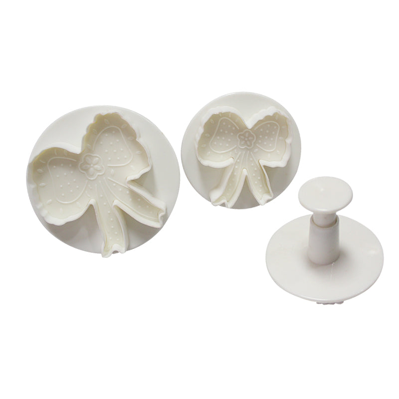 A set of three white plunger cutters in the shape of bow ties, including two bow tie-shaped cutters and one plunger tool. The cutters have decorative details like polka dots and a central flower design.