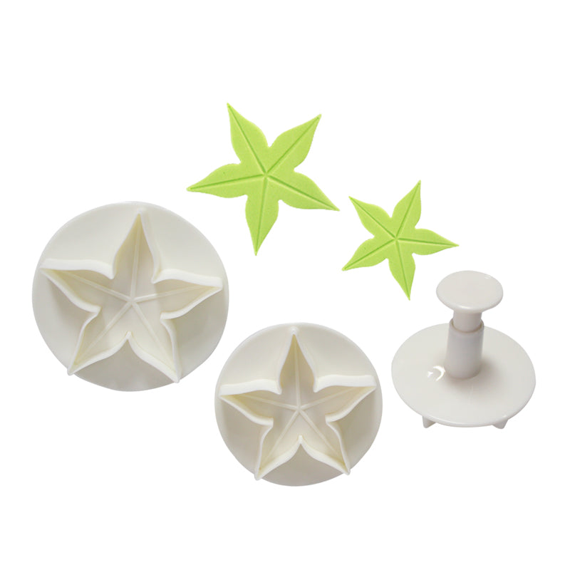 A set of three star-shaped plunger cutters in white plastic. The set includes two larger cutters with multiple star shapes and one smaller plunger cutter for individual stars. There are also two green star-shaped leaves made from an unknown material placed beside the cutters.