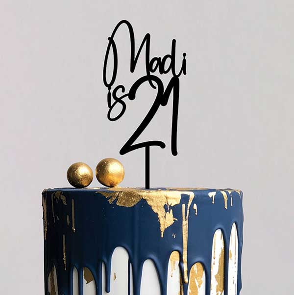 A black acrylic cake topper reading &quot;Madi is 21&quot; is placed on a two-tiered cake. The cake has a navy blue finish with dripping white frosting and gold accents. Two decorative gold spheres sit on top of the cake beside the topper. The background is neutral and simple, focusing on the cake and its decor.