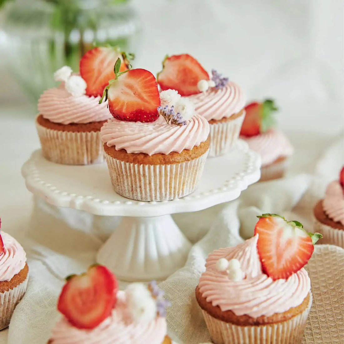 A display of cupcakes with pink frosting, garnished with fresh strawberry slices and small decorative flowers. The cupcakes are arranged on a white cake stand, with additional cupcakes in the background. The setting features soft lighting and light-colored fabrics.