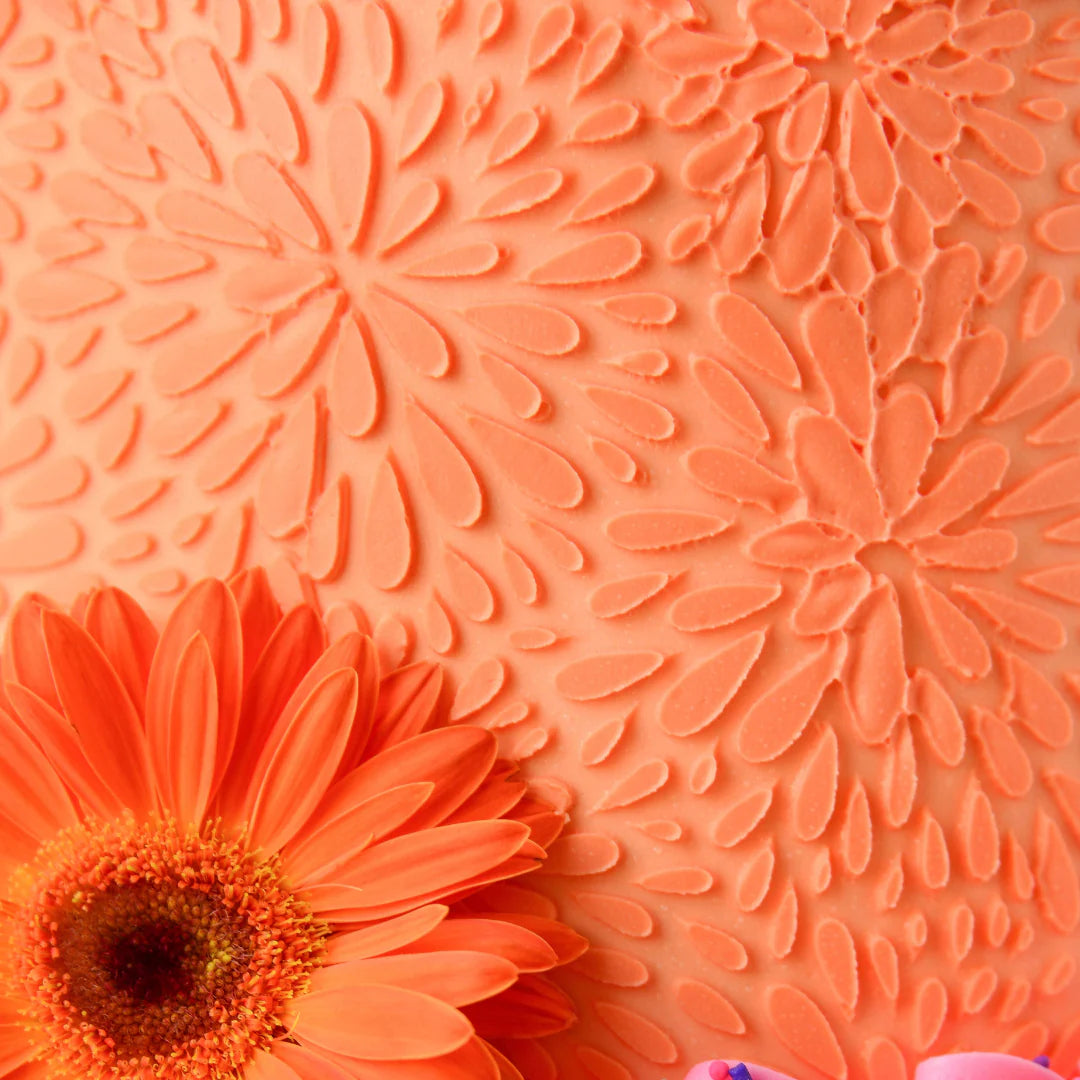 The image features a close-up of a textured surface in a vibrant orange color, resembling decorative icing with floral patterns. A bright orange gerbera daisy is positioned at the bottom left, complementing the background. The overall scene conveys a cheerful and artistic representation suitable for baking or decorating applications.