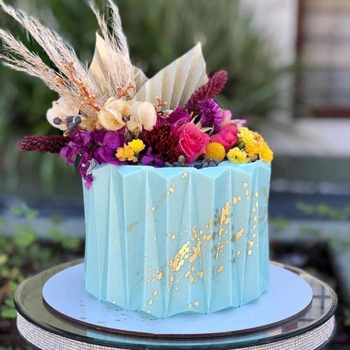 A light blue, origami-style cake with pleated sides, adorned with an arrangement of colorful flowers, including roses and yellow blooms. Gold accents are seen on the cake&