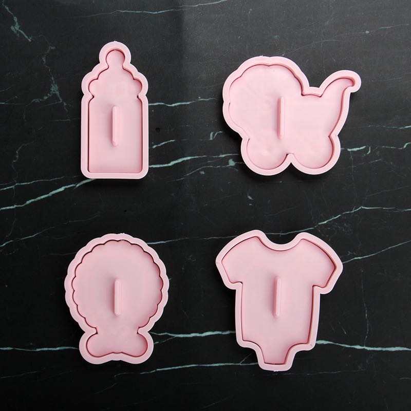 Four pink silicone cutters are arranged on a dark marble surface. The shapes include: 

1. A baby bottle.
2. A stroller.
3. A round object resembling a gift or a decorative item.
4. A baby onesie.

Each cutter features a raised handle for ease of use.