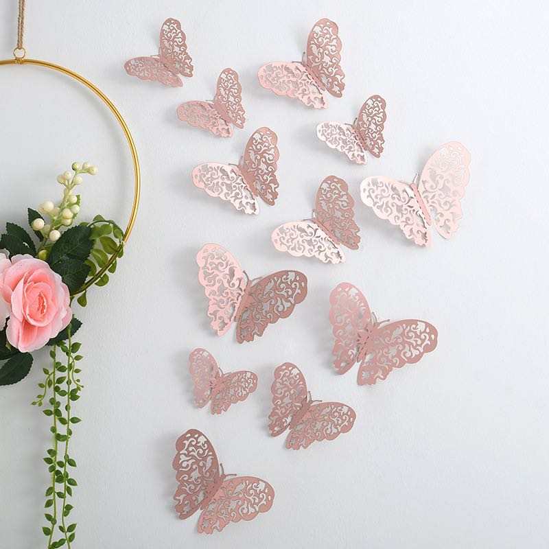 A collection of 12 decorative butterfly stickers in various sizes and designs. The butterflies, displayed on a light-colored wall, feature intricate cut-out patterns and are primarily in shades of silver and light pink. A circular gold hoop with flowers is also partially visible in the image.