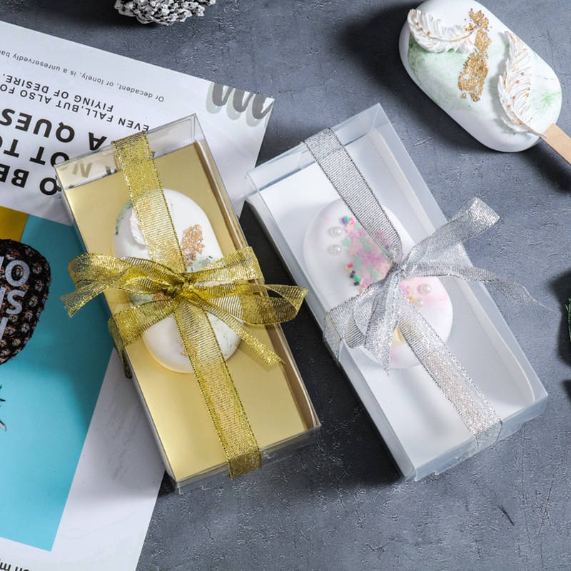 Two elegantly packaged cakesickle boxes are displayed on a textured surface. The left box has a gold ribbon and contains a decorated cakesickle with gold accents. The right box features a silver ribbon and holds a cakesickle adorned with colorful sprinkles. In the background, there are magazines and decorative elements that add to the overall aesthetic.