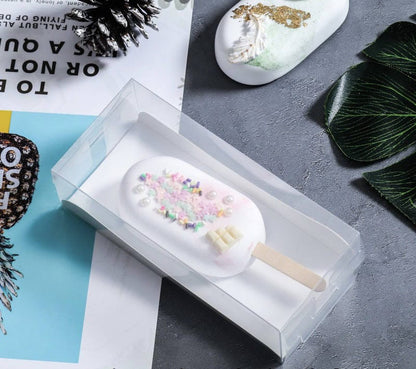 A clear PVC box containing a decorated cakesickle. The cakesickle has a white coating adorned with colorful sprinkles and pearls, and is placed on a wooden stick. The box is displayed on a textured gray surface, alongside a colorful magazine and a leaf.