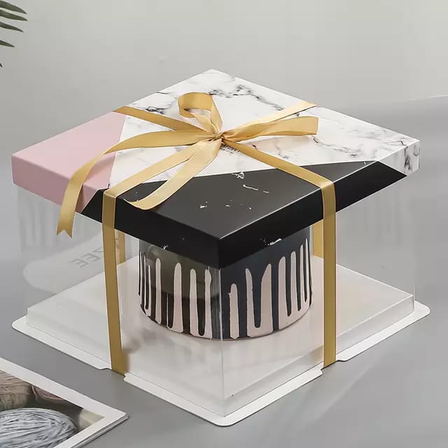 A tall cake box with a square base and a marble-patterned lid is shown. The box features a combination of black, white, and pink colors, with a satin gold ribbon tied around it. Inside, there is a cake with a decorative dripping design. The box is set on a light-colored surface, with some blurred items in the background.