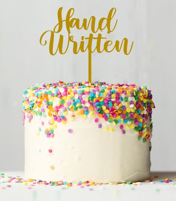 A round cake with white icing and colorful sprinkles on top, featuring a gold cake topper that reads &quot;Hand Written.&quot; The cake is placed on a light background.