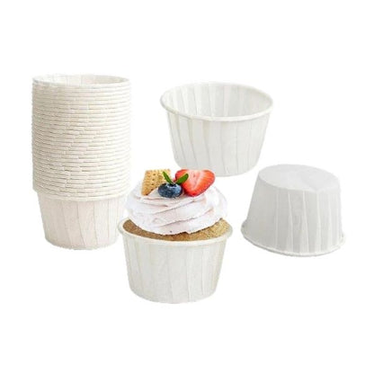 A collection of white paper cupcake liners, including a stack of liners, several individual cups, and one decorated cupcake topped with whipped cream, a strawberry, a blueberry, and a small waffle piece. The liners are fluted and vary in shape and height.