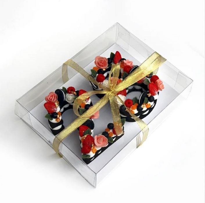 A clear rectangular PVC pastry box contains beautifully decorated pastries in various colors and shapes. The box is elegantly tied with a golden ribbon. Inside, there are pastries featuring a mix of black and white icing, adorned with intricate flower decorations in shades of pink, red, and green, along with small colorful fruit-like accents. The overall presentation is visually appealing and suggests a festive or celebratory occasion.