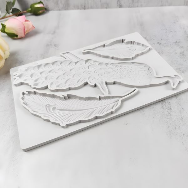 A white silicone mould featuring a peacock and two feather shapes, designed for crafting or baking. The mould is flat and includes intricate detailing of the peacock and feathers. In the background, there are soft pink roses and a light-colored surface.