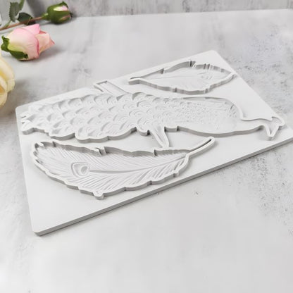 A white silicone mould featuring a peacock and two feather shapes, designed for crafting or baking. The mould is flat and includes intricate detailing of the peacock and feathers. In the background, there are soft pink roses and a light-colored surface.
